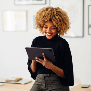 Cheerful African American woman, sitting on her desk, smiling and using her laptop. She is typing something.