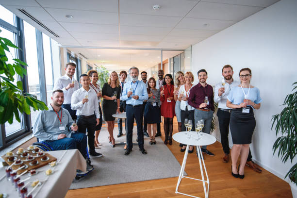 Group portrait of bearded CEO in mid 50s standing with staff of new business and holding glass of sparkling wine for celebratory toast.